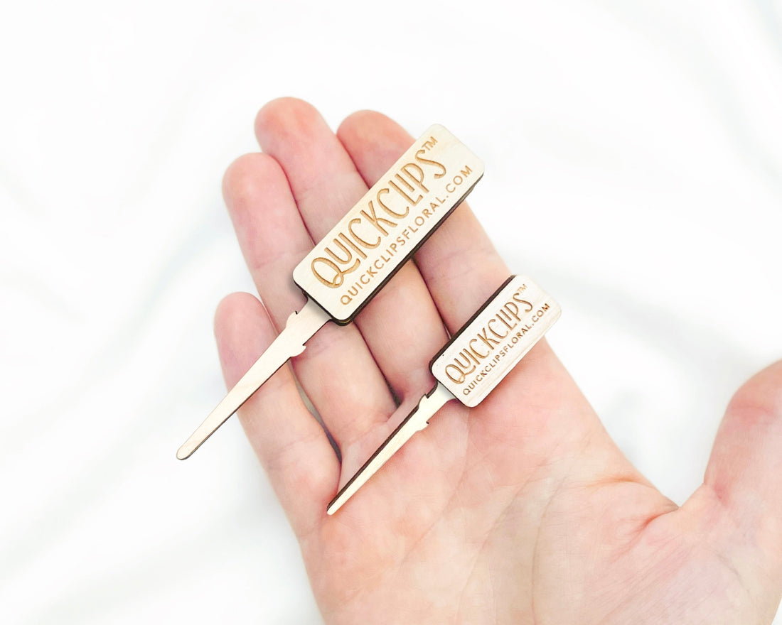 A comparison of a QuickClip Original and a QuickClip Mini on a hand above a white fabric background